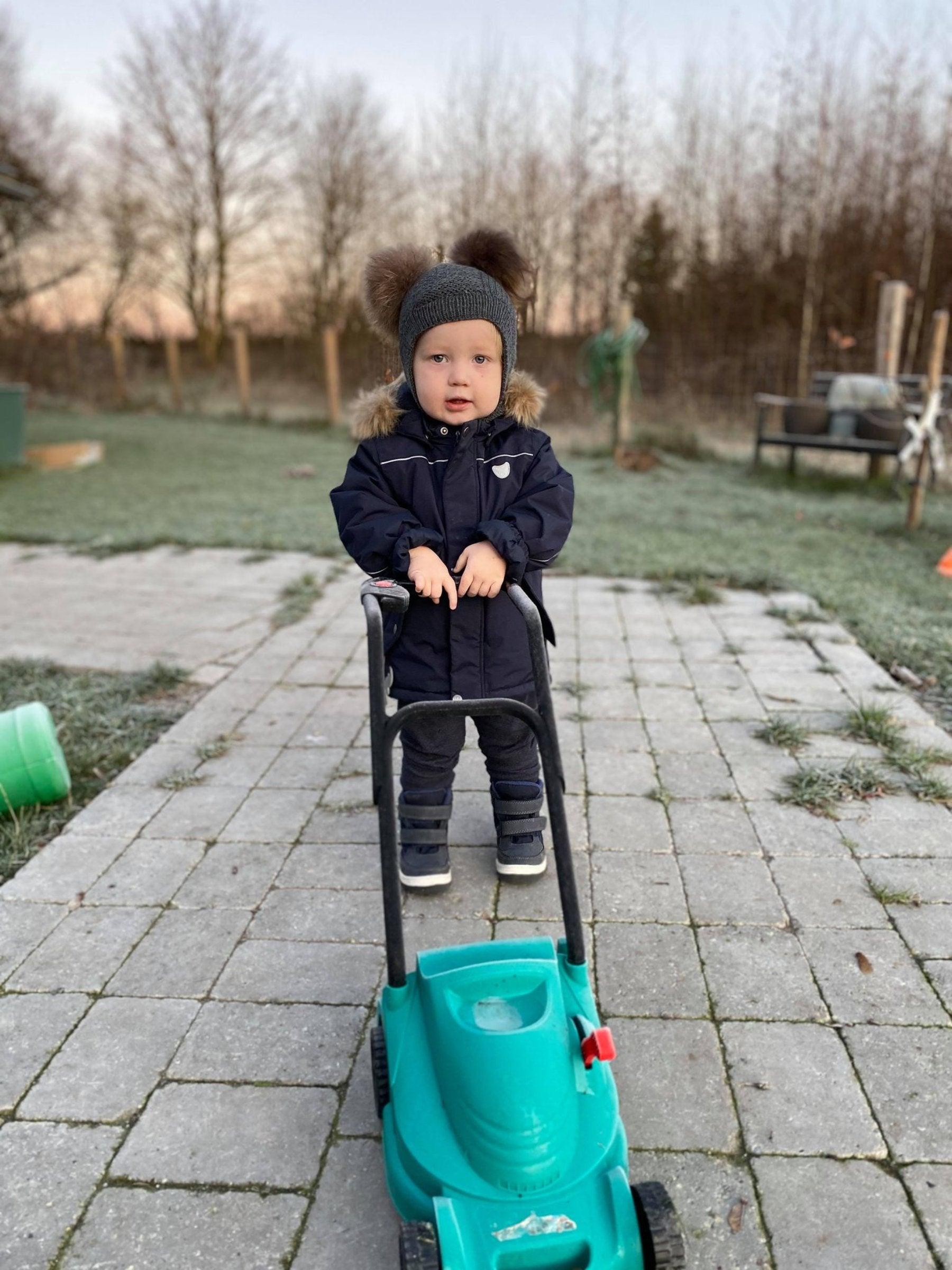 Lille barn i mørkeblå flyverdragt udendørs på en frostklar terrasse med grå strikhue med to store kvaste